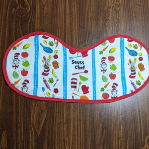 May include: A white and red pot holder with a Seuss Chef theme. The pot holder features a repeating pattern of kitchen utensils, vegetables, and the Cat in the Hat. The text "Seuss Chef" is printed in blue on the pot holder.