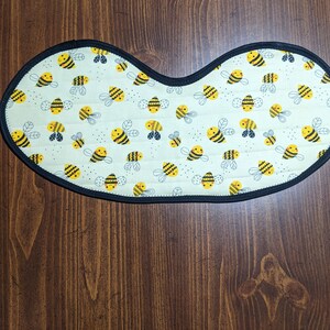 May include: A white fabric placemat with a black border and a repeating pattern of yellow and black bees.