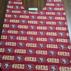 May include: Red fabric bib with a repeating pattern of the San Francisco 49ers logo. The bib is labeled "MEDIUM".