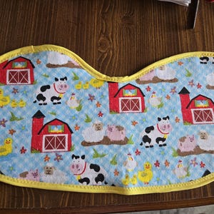 May include: A farm-themed baby bib with a blue gingham background. The bib features red barns, black and white cows, yellow chicks, pink pigs, and white sheep. The bib is trimmed with yellow fabric.