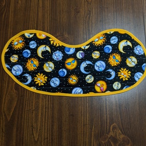 May include: A black fabric trivet with a yellow border features a repeating pattern of suns and moons with faces wearing sunglasses. The background is black with white polka dots.