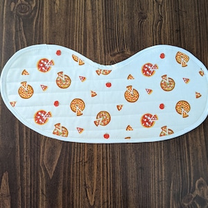 May include: White fabric baby bib with a pizza pattern. The bib has a rounded shape and is quilted. The pizza pattern includes red, yellow, and brown pizza slices, white cheese, and red tomato slices.