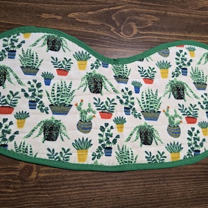 Peut inclure: Un bavoir blanc en forme de bib, bordé de vert. Le bavoir est orné de diverses plantes en pot dans des tons de vert, bleu, jaune et orange. Le bavoir est probablement en tissu doux.