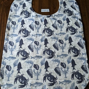 May include: A large, cream-colored bib with a repeating pattern of blue crabs, fish, lobster, and shells. The bib has a curved neckline and a Velcro closure. The word "LARGE" is printed on the bib.