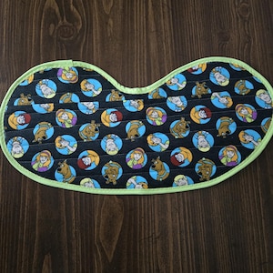 Op de afbeelding: Een zwart, ovaalvormig stoffen item met een groene rand. De stof is bedrukt met stripfiguren in blauwe cirkels op een zwarte achtergrond. De figuren zijn onder meer een hond en een blonde vrouw.