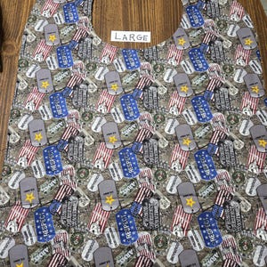 May include: A large, patterned baby bib featuring a repeating design of military dog tags, American flags, and stars. The bib has a neck opening and is labeled "LARGE". The color palette includes shades of gray, blue, red, and yellow.