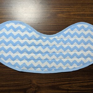 May include: A light blue baby bib with a white chevron pattern. The bib is made of soft, quilted fabric and has a rounded shape.