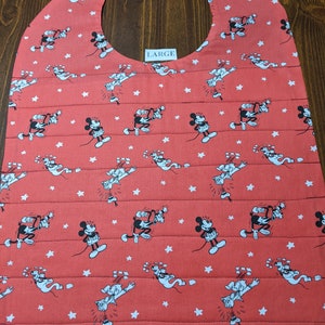 Puede incluir: Un babero rojo con un patrón de estrellas blancas y Mickey Mouse. El babero tiene una abertura grande para el cuello y está etiquetado como "LARGE".