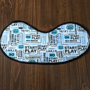 May include: A white eye mask with a black border. The mask is covered in a repeating pattern of blue and black text that says "Start Play", "Score", "Level", "Champion", "Let's Go!" and "Bonus Game".