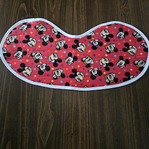 Op de afbeelding: Rode, gewatteerde baby slabbetje met een witte rand. Het slabbetje heeft een herhalend patroon van Mickey Mouse gezichten en kleurrijke sterren. Ontworpen om gemors op te vangen en kleding te beschermen.