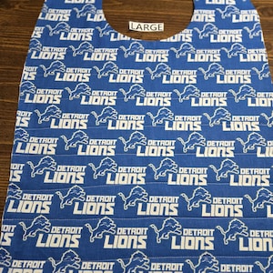 May include: A blue fabric bib featuring a repeating white pattern of the Detroit Lions logo and text. The bib has a rounded neck opening and a small white rectangular tag near the top center with the word LARGE printed in black text.