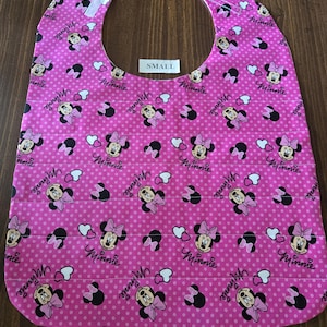 May include: Pink fabric baby bib with a Minnie Mouse print. The bib has white polka dots and the text "SMALL" on a white tag.