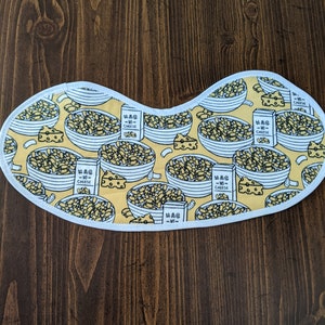 Puede incluir: Una máscara de dormir blanca con fondo amarillo que presenta un patrón repetido de cuencos de macarrones con queso y cajas de macarrones con queso. El texto en las cajas dice "Mac & Cheese".