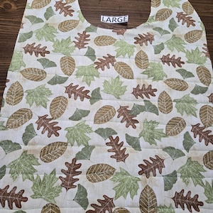 May include: A large, quilted bib with a cream background and a repeating pattern of brown and green leaves. The word "LARGE" is printed on a white label. The bib has a curved neckline.