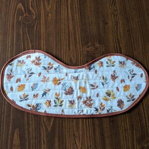 May include: A white quilted placemat with a brown trim and a pattern of autumn leaves in shades of orange, yellow, and brown.