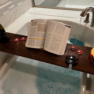 May include: A wooden bath tray with a book, a glass of wine, and candles. The tray is placed over a bathtub filled with water and bubbles.