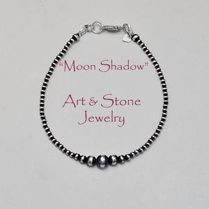 May include: A silver bracelet with black and silver beads. The bracelet has a heart-shaped charm near the clasp. The text "Moon Shadow" and "Art & Stone Jewelry" are in the image.