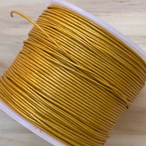 May include: A spool of gold-colored leather cord. The cord is wound tightly around the spool, which is made of white plastic.