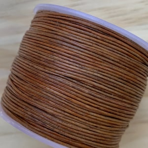 May include: A spool of brown leather cord, wound tightly in a spiral shape. The cord is smooth and has a natural, rustic look.