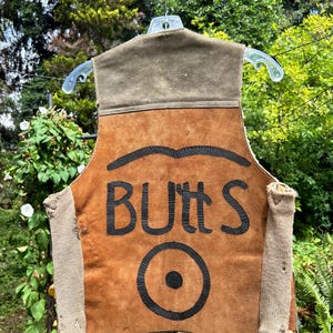 May include: A sleeveless vest made of tan suede leather with a dark olive green upper panel. The back features the black painted text "BUTTS" above a black circular graphic. The sides are constructed from a textured, light beige fabric material.
