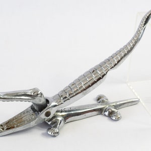 May include: A chrome-plated nutcracker in the shape of an alligator. The nutcracker features a detailed design with textured scales and a hinged jaw. The alligator's tail curves upwards, and its legs are visible at the base.