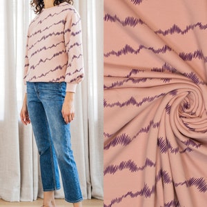 Könnte beinhalten: Ein rosa Sweatshirt mit einem violetten, wellenförmigen Muster. Das Sweatshirt hat einen Rundhalsausschnitt und lange Ärmel. Das Model trägt eine blaue Jeans und violette Federsandalen. #ruby
