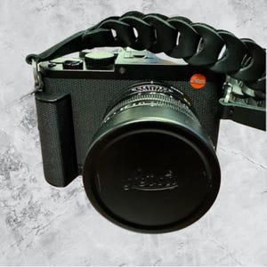 Op de afbeelding: Zwarte camera met een lensdop en een gevlochten zwarte leren riem. De camera heeft een getextureerde behuizing en een rood logo. De lens is bedekt met een zwarte dop met de merknaam.