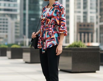 May include: A kimono-style jacket featuring a vibrant abstract pattern in shades of red, blue, white, and black, with three-quarter length sleeves and a matching fabric tie belt at the waist, worn over a white top with black trousers and loafers.