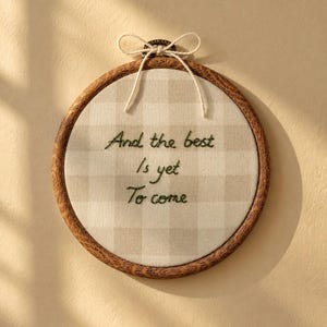 Könnte beinhalten: Ein runder Stickrahmen mit einem hellbeigen und weißen karierten Stoff. Der Text 'And the best Is yet To come' ist mit dunkelgrünem Garn in die Mitte gestickt. Eine dünne weiße Schnur ist oben am Rahmen zu einer Schleife gebunden.