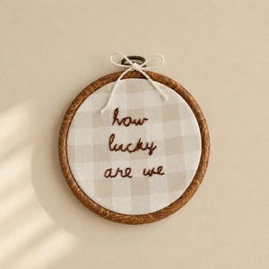 May include: A circular wooden embroidery hoop framing a light beige and white checkered fabric. The fabric features the hand-stitched brown text "how lucky are we" centred in the middle. A thin white string is tied in a bow at the top metal screw.