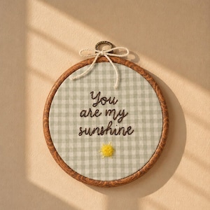 May include: A circular embroidery hoop with a wood-grain frame containing light green and white gingham fabric. The text "You are my sunshine" is stitched in dark brown thread, accompanied by a small, textured yellow sun embroidery below.
