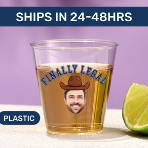 May include: A clear plastic shot glass filled with a golden liquid. The cup features a graphic of a person wearing a brown cowboy hat with the text FINALLY LEGAL printed in blue arched lettering above the face. A lime wedge sits to the right.