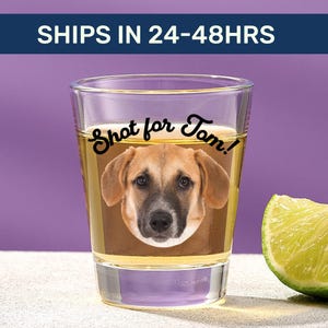 May include: A clear glass shot glass filled with a golden liquid, featuring a printed image of a brown dog face and the black text Shot for Tom! positioned above the dog. A fresh green lime wedge sits to the right of the glass.