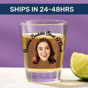 May include: A clear glass shot glass filled with a golden liquid, featuring a printed image of a person's face and the black text Baddie Baddie Shot O' Clock. A fresh lime wedge sits to the right of the glass on a light grey surface.