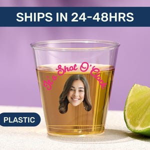 May include: A clear plastic shot glass filled with a golden liquid, featuring a woman's face and the text "It's Shot O'Clock" in pink. A lime wedge rests beside the glass. The word "PLASTIC" is in a blue rectangle.