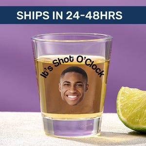 May include: A clear glass shot glass filled with a golden liquid, featuring a printed photograph of a smiling person's face and the black text "It's Shot O'Clock" arched above the face. A fresh lime wedge sits beside the glass.