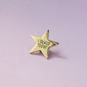 Puede incluir: Un pin dorado en forma de estrella con el texto "I TALKED TODAY" en letras negras. El pin está sobre un fondo lila suave. Un accesorio sencillo pero llamativo.