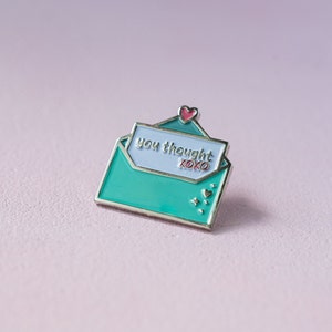 Puede incluir: Un pin de esmalte turquesa con forma de sobre abierto. El pin presenta las palabras "you thought" y "xoxo" en una tarjeta azul claro. Un pequeño corazón rosa se encuentra encima de la tarjeta, con detalles de estrellas y corazones.