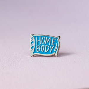 Puede incluir: Un pin de esmalte azul con forma de almohada con las palabras "HOME BODY" en letras blancas. El pin tiene un borde plateado y está sobre un fondo rosa claro.