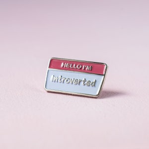Puede incluir: Un pin rectangular de esmalte con borde plateado. La sección superior es roja con las palabras "HELLO I'M" en plata. La sección inferior es blanca con la palabra "Introverted" en plata. El pin está sobre un fondo rosa.