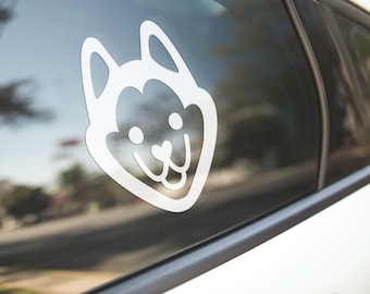 Pode incluir: Um autocolante de vinil branco com o rosto de um cão, aplicado no vidro de um carro. O design apresenta uma representação simplificada, em estilo cartoon, da cabeça de um cão com orelhas grandes, olhos redondos e um nariz em forma de coração. O autocolante é limpo e moderno.