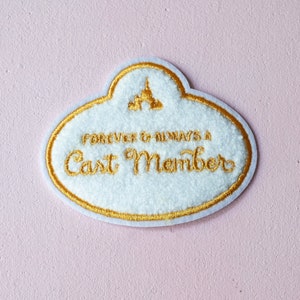 May include: White and gold embroidered patch with the text "FOREVER & ALWAYS A Cast Member" and a castle graphic. The patch has a gold border and is on a pink background.