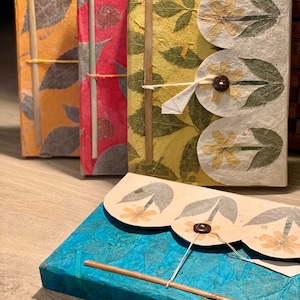 Puede incluir: Colección de diarios y cuadernos hechos a mano. Las cubiertas presentan hojas y flores prensadas en varios colores, incluyendo naranja, rojo, amarillo y azul. Cada diario está encuadernado con un palito de madera y un cierre de cuerda.
