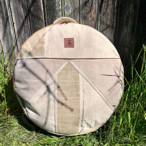 Peut inclure: Un sac rond en toile beige avec une sangle en cuir marron et un petit patch en cuir marron avec un logo. Le sac est conçu pour transporter un tambour.
