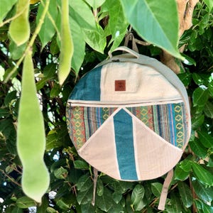 Peut inclure: Sac à dos rond avec un design beige, turquoise et à motifs. Il possède une poignée supérieure, une fermeture éclair et des bretelles réglables. Le sac est suspendu à une branche d'arbre avec des feuilles vertes et de longues gousses vertes.