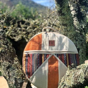 Peut inclure: Un sac de tambour rond en tissu blanc, orange et multicolore avec une fermeture éclair et une poignée en cuir marron. Le sac est suspendu à une branche d'arbre.