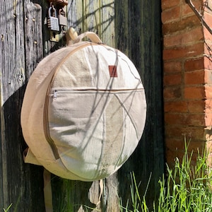 Pode incluir: Uma bolsa de bateria de lona branca com um bolso com zíper e alças ajustáveis. A bolsa está pendurada em uma porta de madeira.
