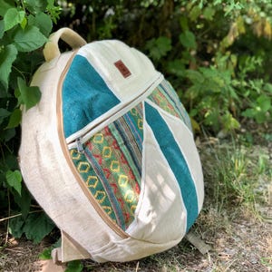 Op de afbeelding: Een beige rugzak met turquoise en patroonaccenten. De rugzak heeft een voorvak met ritssluiting en een handvat aan de bovenkant. Het ontwerp bevat een mix van texturen en kleuren, met een bruin leren label.