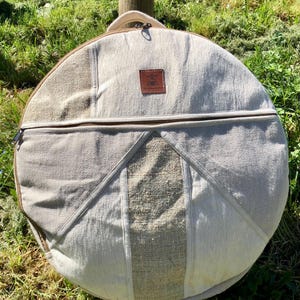 Peut inclure: Un sac rond beige avec une fermeture éclair et une poignée. Le sac est fait d'un tissu texturé avec un patch en cuir marron. Le sac est posé sur de l'herbe verte.
