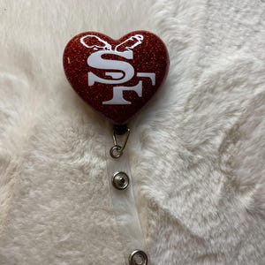 Peut inclure: Un porte-badge en forme de cœur rouge pailleté avec les lettres blanches "SF". Le porte-badge est doté d'un cordon rétractable en plastique transparent et d'un clip en métal. Le fond est une surface blanche et duveteuse.